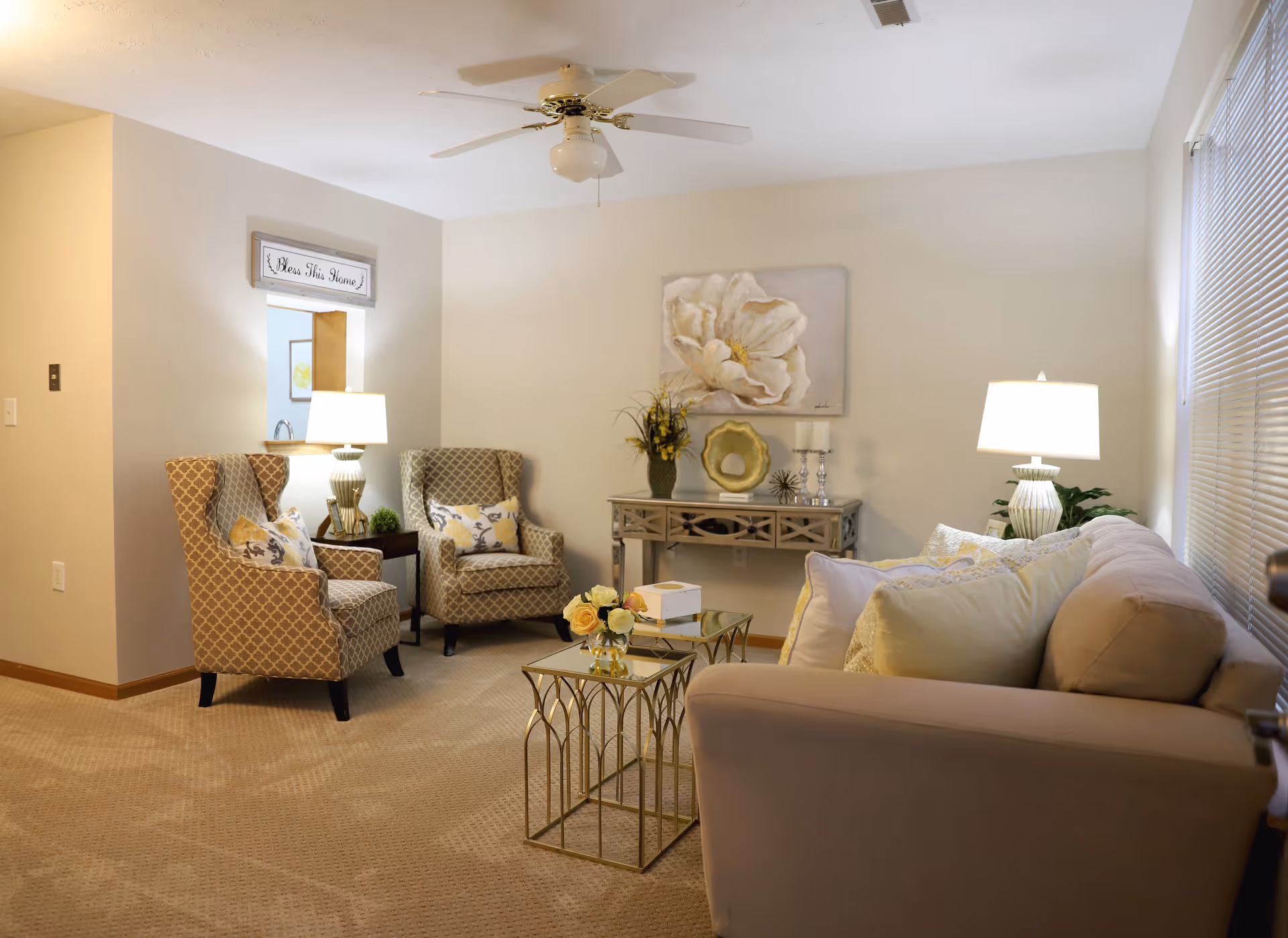 A cozy living room with a beige sofa adorned with multiple pillows, two patterned armchairs with decorative cushions, two side tables with lamps, a glass coffee table with a flower arrangement, a console table with decorative items, and a large floral painting on the wall. The room has beige carpeting and neutral-colored walls.