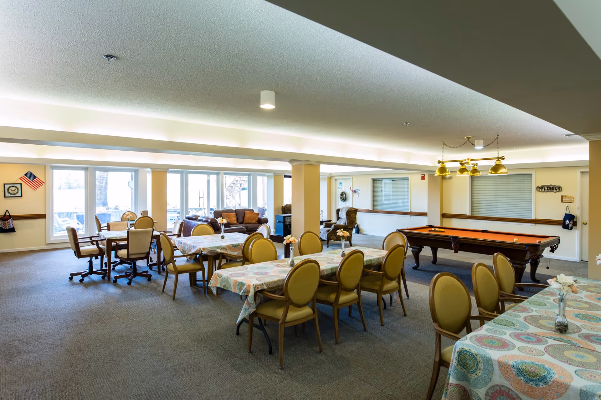 A spacious common area in a senior living facility featuring multiple tables with patterned tablecloths and chairs arranged around them. In the background, there is a pool table with overhead lighting and a seating area with sofas near large windows letting in natural light. The room has a carpeted floor and neutral-colored walls.