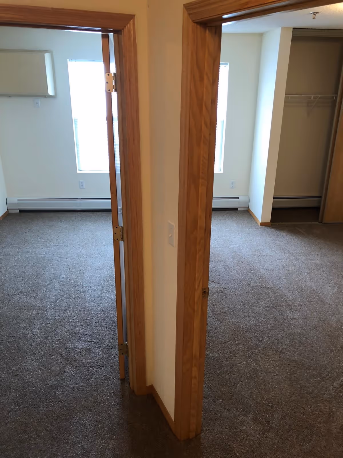 Carpeted interior showing two open doorways into empty bedrooms with windows, baseboard heaters, and a closet.