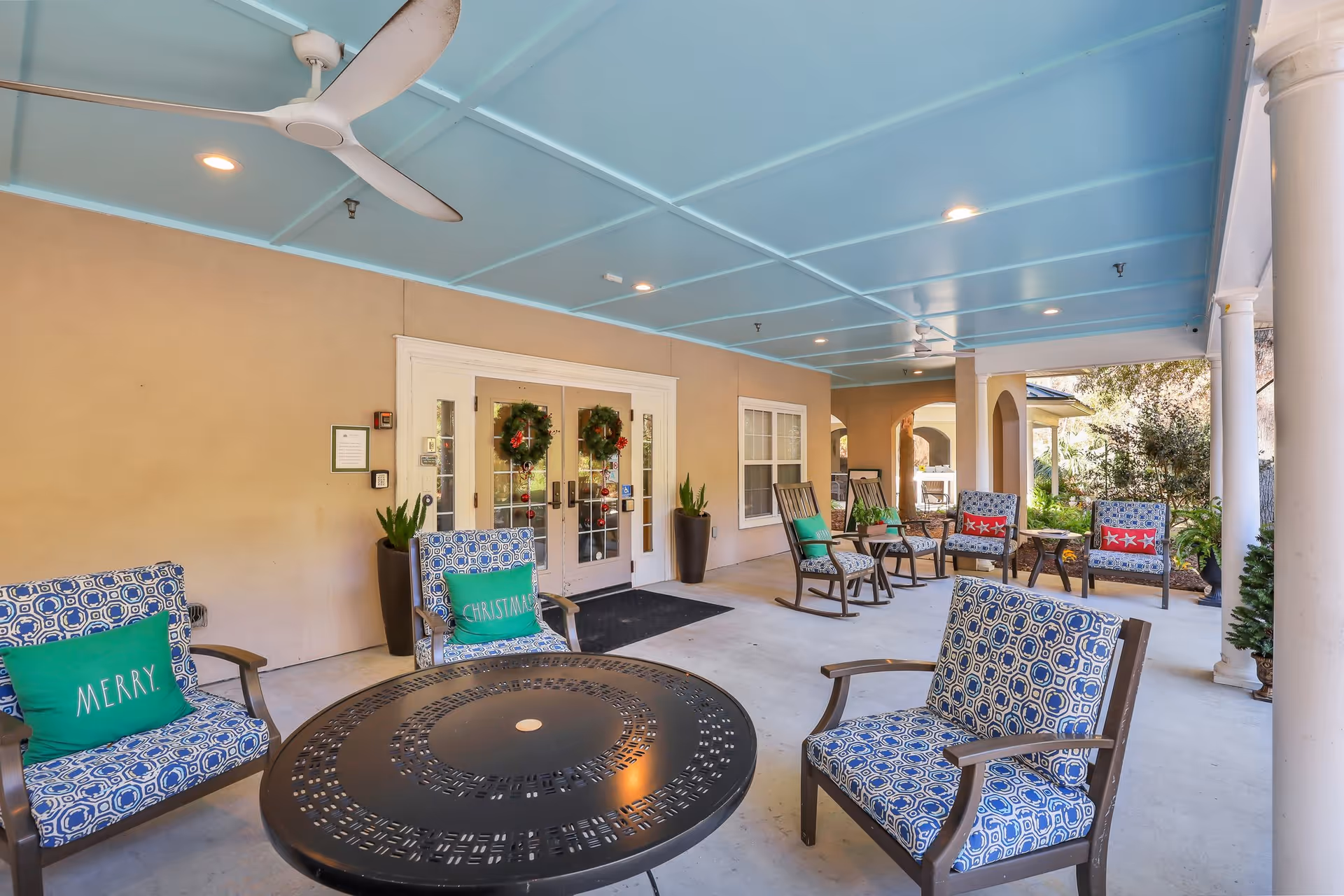 Covered outdoor patio area with blue ceiling, ceiling fans, and multiple cushioned chairs arranged around a round black metal table. The chairs have patterned blue cushions and some have green or red holiday-themed pillows. Double doors decorated with Christmas wreaths lead inside the building. Large potted plants flank the doors.