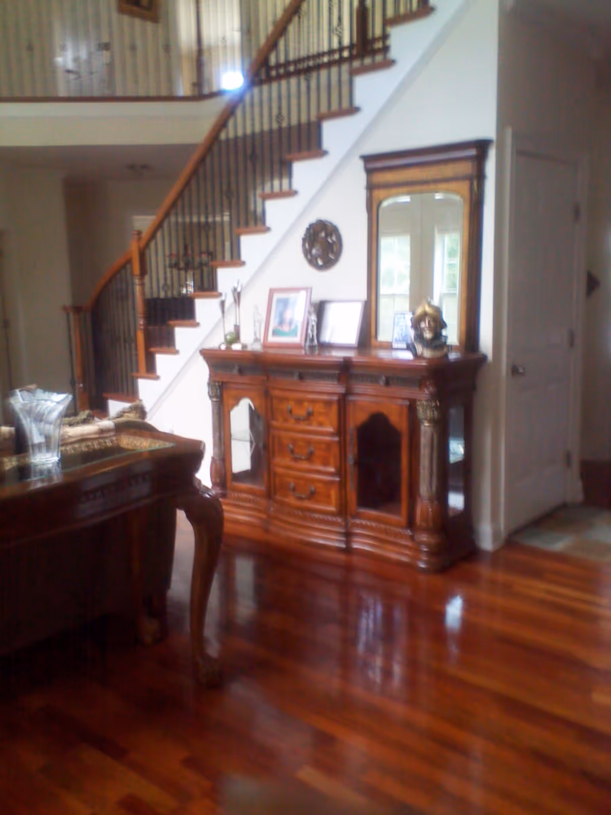 A polished wood-floored living area with a curved staircase and an ornate wooden sideboard topped with a mirror and decorative items.