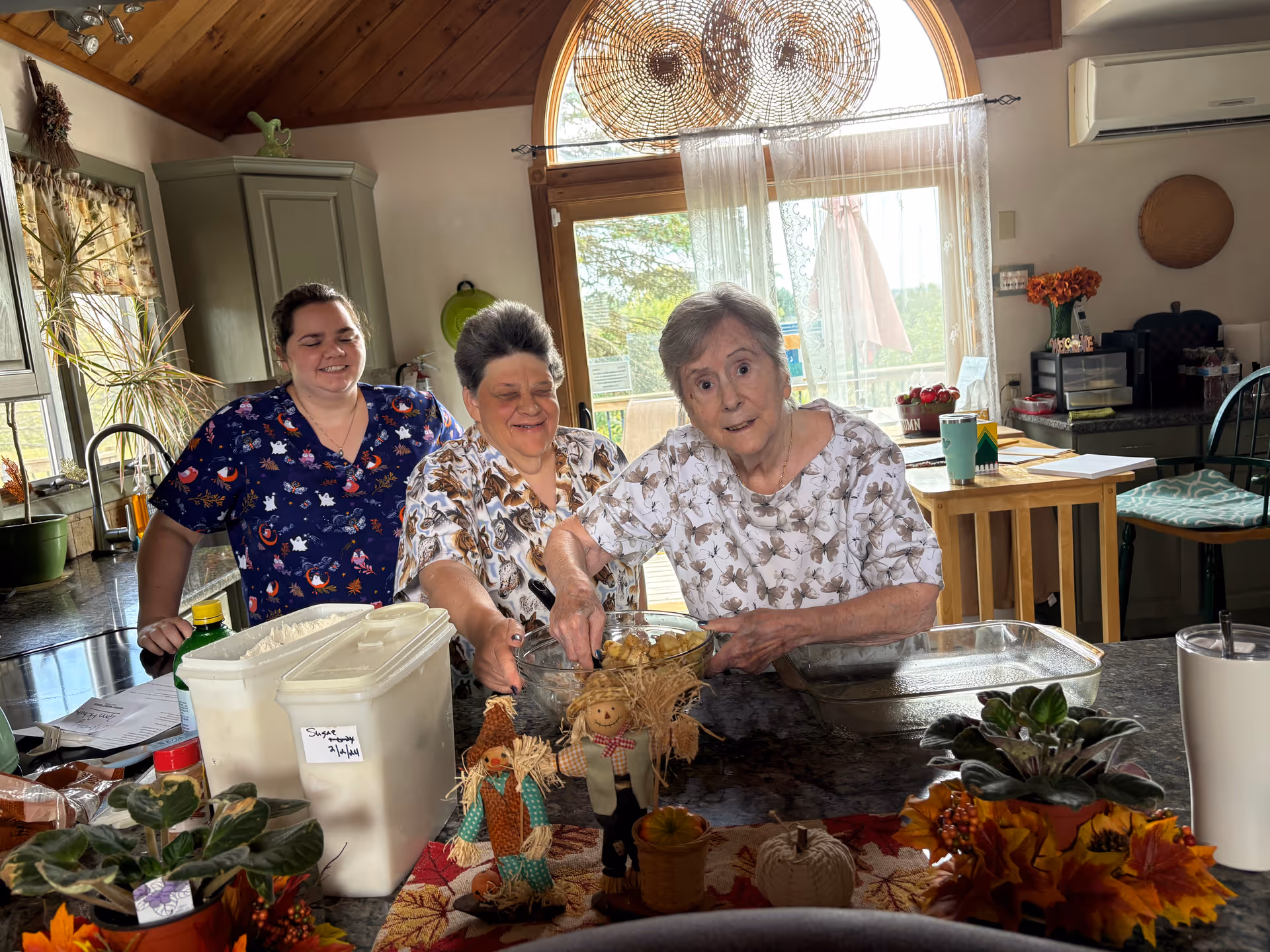 Three women in a kitchen preparing food together. Two elderly women and a younger woman are smiling and mixing ingredients in a glass bowl on a kitchen counter. The kitchen has wooden cabinets, a large window with sheer curtains, and various decorative items including small scarecrow figurines and autumn-themed decorations on the counter.