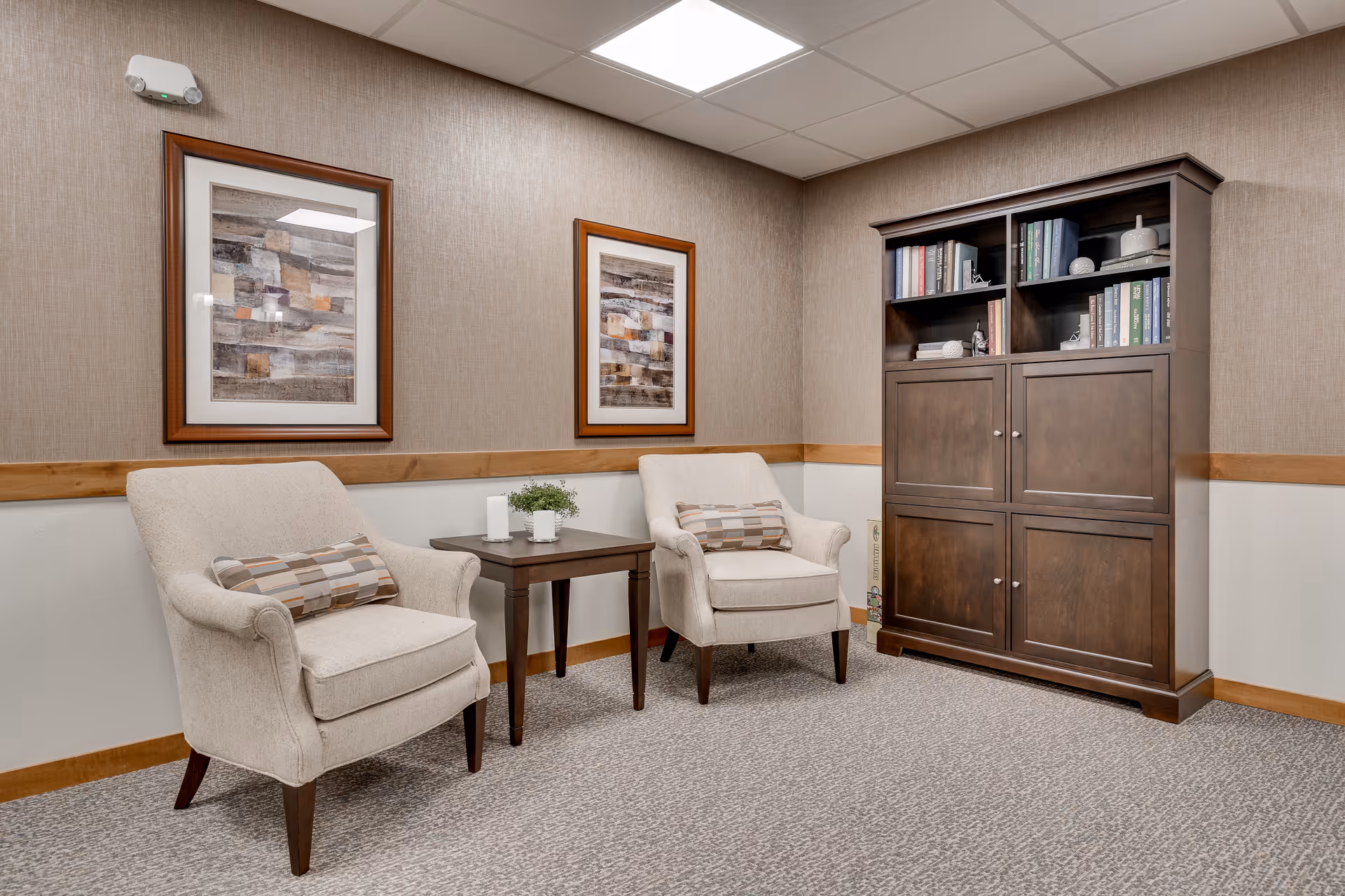 A small seating area with two upholstered armchairs, a side table, framed artwork, and a wooden cabinet in a senior living facility.