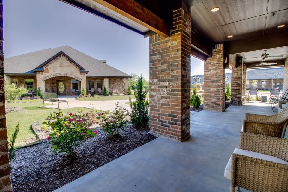 Covered outdoor patio area with brick columns and wicker chairs, overlooking a landscaped garden with flowering bushes and a brick building with a peaked roof in the background under a clear blue sky.