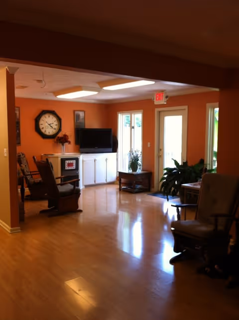 A cozy living room area with orange walls and wooden flooring. The room features several cushioned rocking chairs, a white cabinet with a flat-screen TV on top, a large wall clock, and a small table with a plant near a glass door leading outside. The room is well-lit with ceiling lights and natural light coming through the windows.