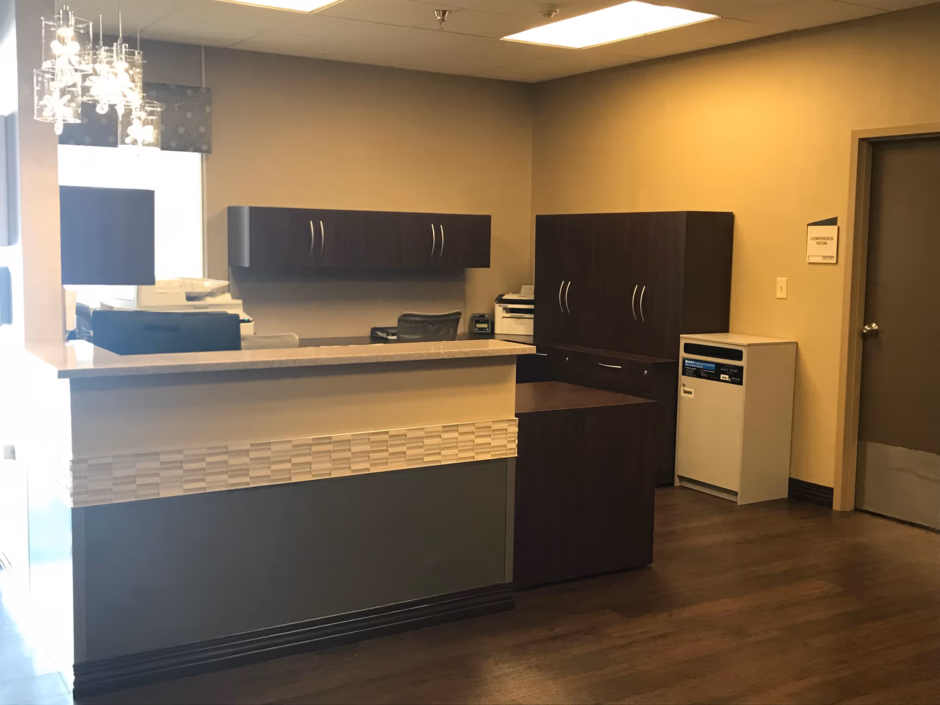 Reception area with a front desk, dark wood cabinets, office chair, printer, and a door labeled 'Conference Room' in the background. The room has beige walls and wood flooring.