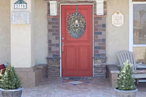 Red front door with wreath, stone porch, and outdoor seating beside entrance.
