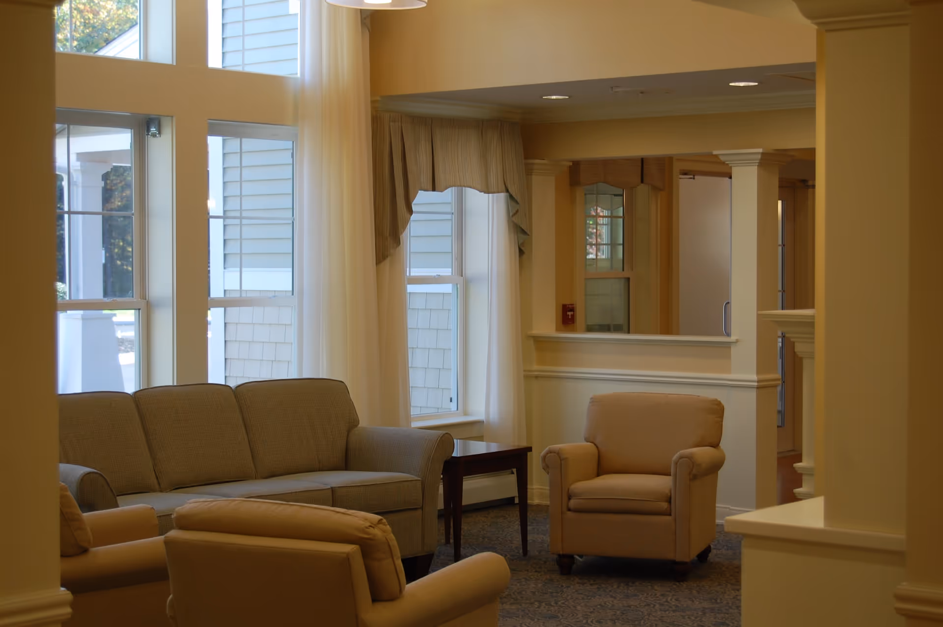 A cozy living room area with beige armchairs and a sofa arranged around a small wooden side table. Large windows with white curtains allow natural light to fill the space. The walls are painted in a soft yellow tone, and there is a carpeted floor with a subtle pattern.