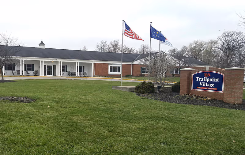Exterior view of Trailpoint Village senior living community building with a large green lawn in front, three flagpoles flying the American flag, a state flag, and another flag, and a brick sign displaying the facility name.