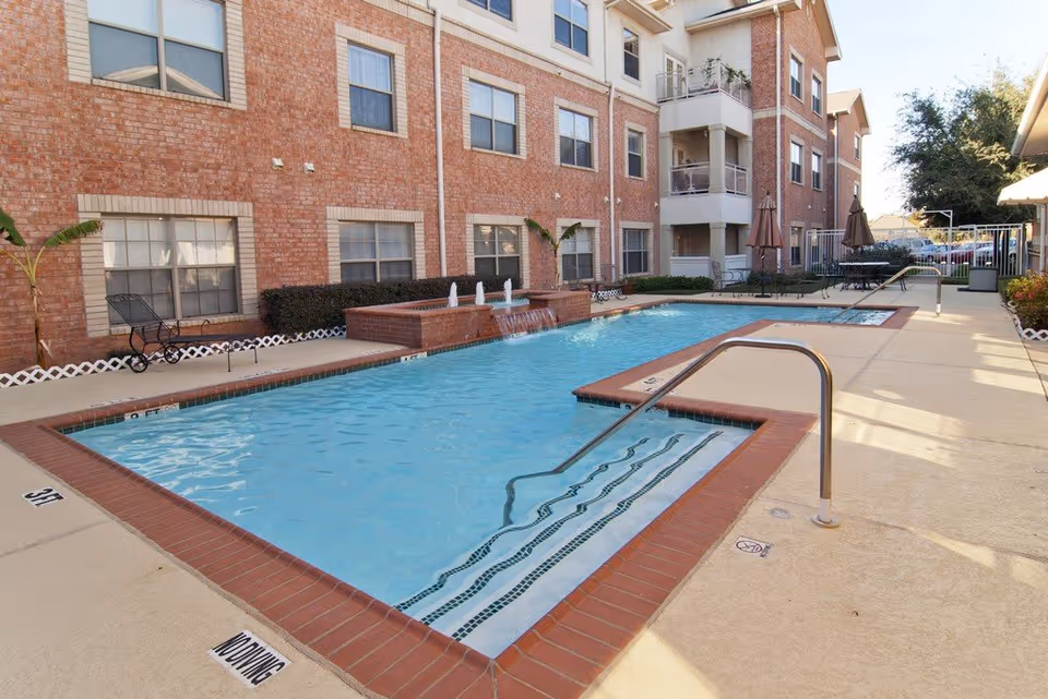 Outdoor swimming pool area at The Remington at Valley Ranch with clear blue water, surrounded by a beige concrete deck. The pool has metal handrails and steps leading into the water. There are brick buildings with multiple windows in the background, along with patio tables, chairs, and umbrellas near the pool. Some small plants and bushes are also visible around the pool area.