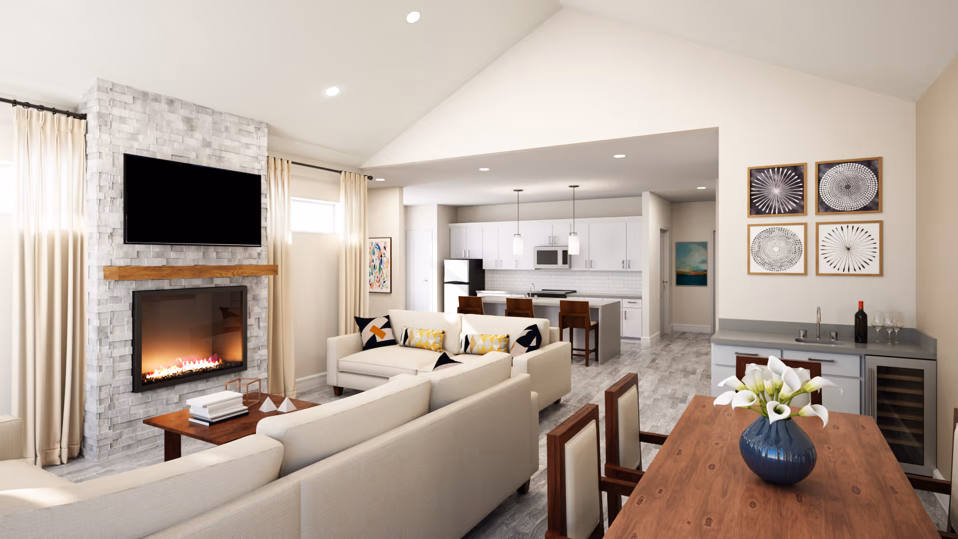 A bright and modern living room with a stone fireplace and a mounted flat-screen TV above it. The room features two beige sofas with decorative pillows, a wooden coffee table with books and decor, and large windows with cream curtains. In the background, there is an open kitchen with white cabinets, a refrigerator, a microwave, and a kitchen island with three bar stools. To the right, there is a dining area with a wooden table, chairs, a blue vase with white flowers, and a small counter with a wine cooler and wine glasses. Four framed abstract artworks hang on the wall above the counter.