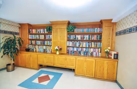 Wooden bookshelves line a warm library area with plants and decorative floor tiles.