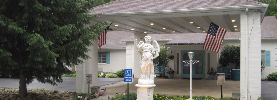 Covered entrance of a senior living building with a white statue, American flags, and double doors under a porte-cochere.