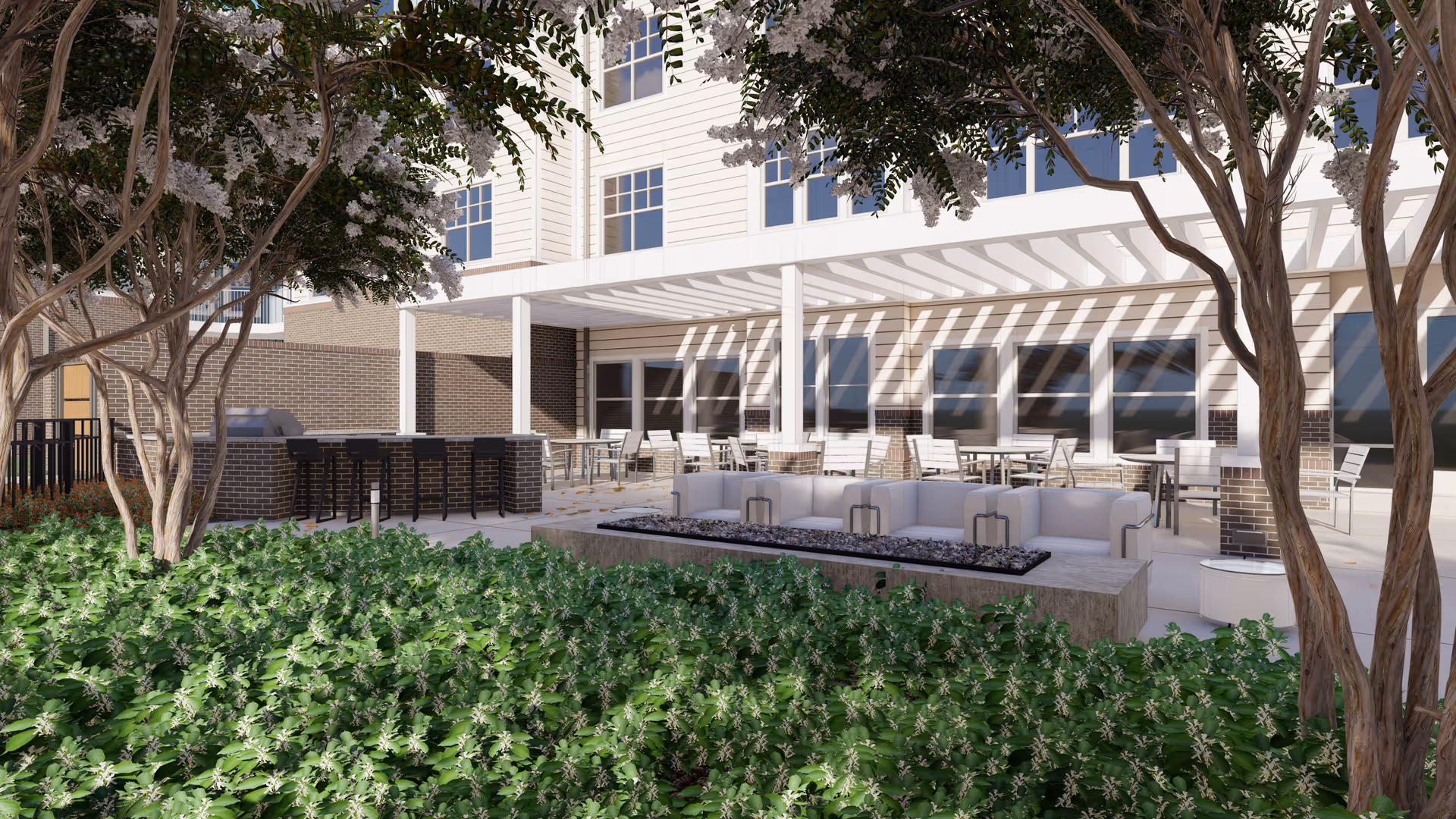 Outdoor patio area at a senior living facility featuring a covered seating area with tables and chairs, a bar with stools, a fire pit surrounded by cushioned seating, and lush greenery with trees and bushes in the foreground.