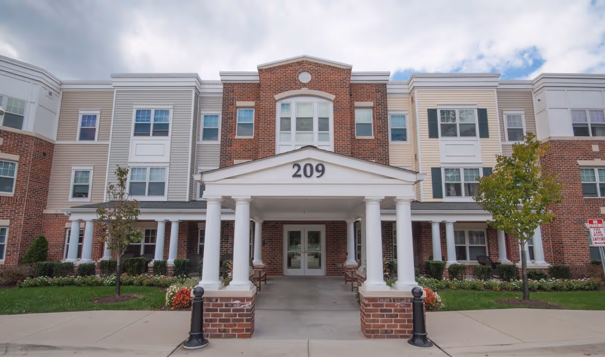 Front exterior view of a senior living facility building with a covered entrance supported by white columns, the number 209 displayed above the entrance, brick and beige siding, multiple windows, and landscaped greenery with small trees and flowers.