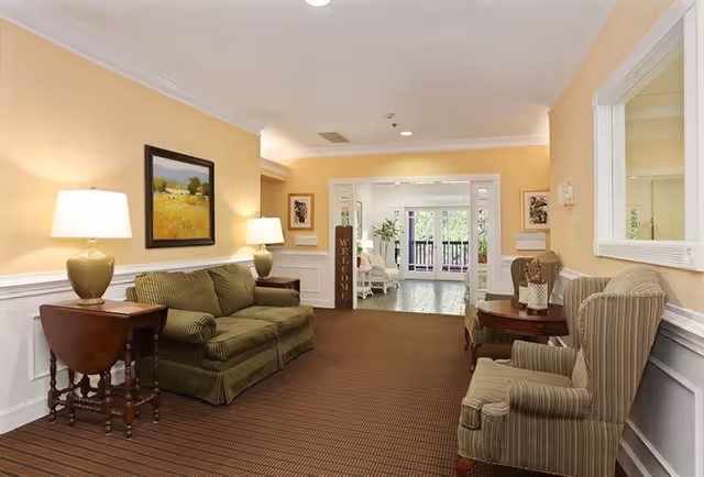 A cozy living room area in a senior living facility with beige walls and white trim. The room features a green striped sofa with two matching table lamps on wooden side tables, a round wooden table with two striped armchairs, a large mirror on the wall, and a welcome sign near the entrance to a sunlit porch with white wicker furniture and greenery outside.