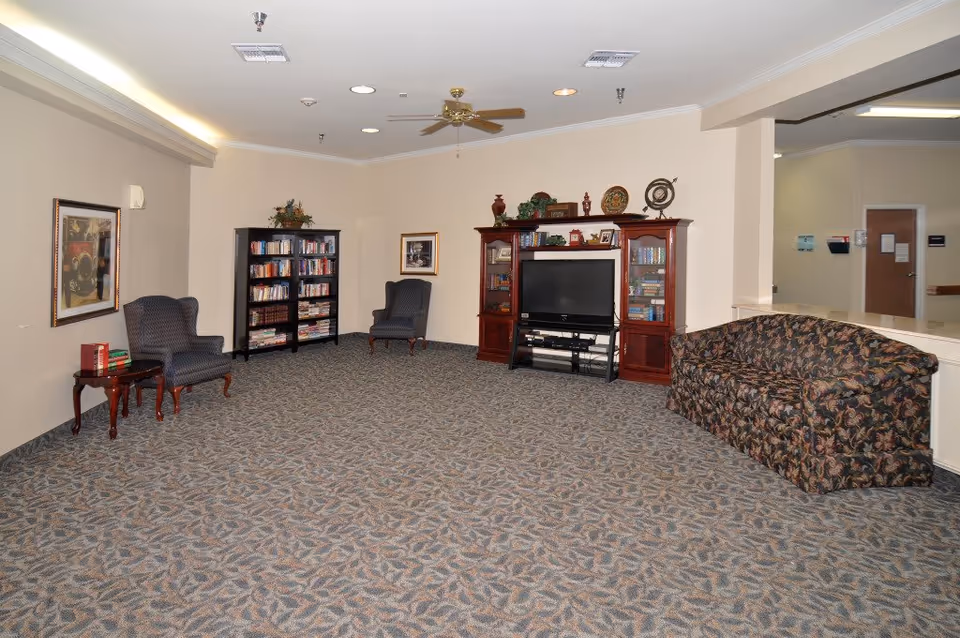 A common area in a senior living facility with patterned carpet flooring, two upholstered armchairs with a small wooden side table holding books, a bookshelf filled with books, a wooden entertainment center with a flat-screen TV and decorative items, and a floral-patterned couch. The walls are beige with framed artwork, and there is a ceiling fan and recessed lighting.