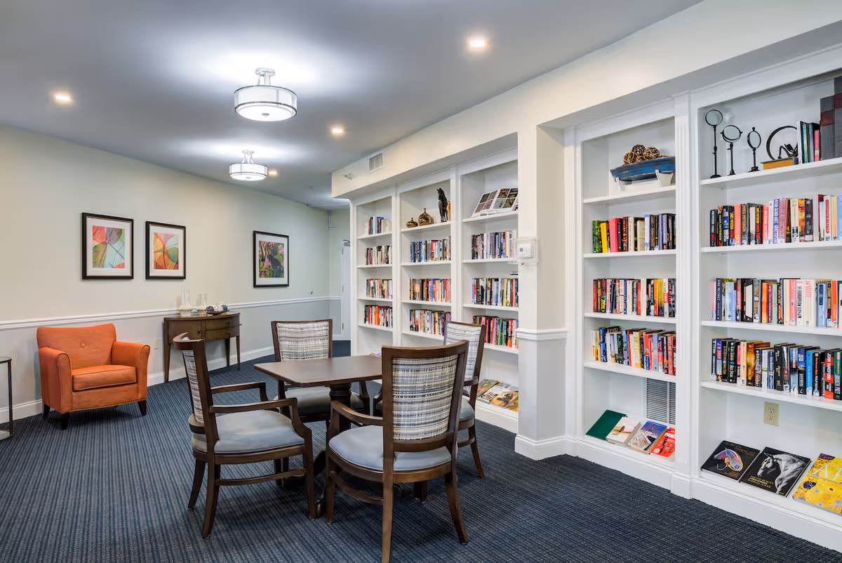 A cozy reading and sitting area in a senior living facility featuring a wooden table with four cushioned chairs, white built-in bookshelves filled with books and decorative items, an orange armchair, a small wooden side table, and three framed abstract artworks on the wall. The room has carpeted flooring and ceiling lights.