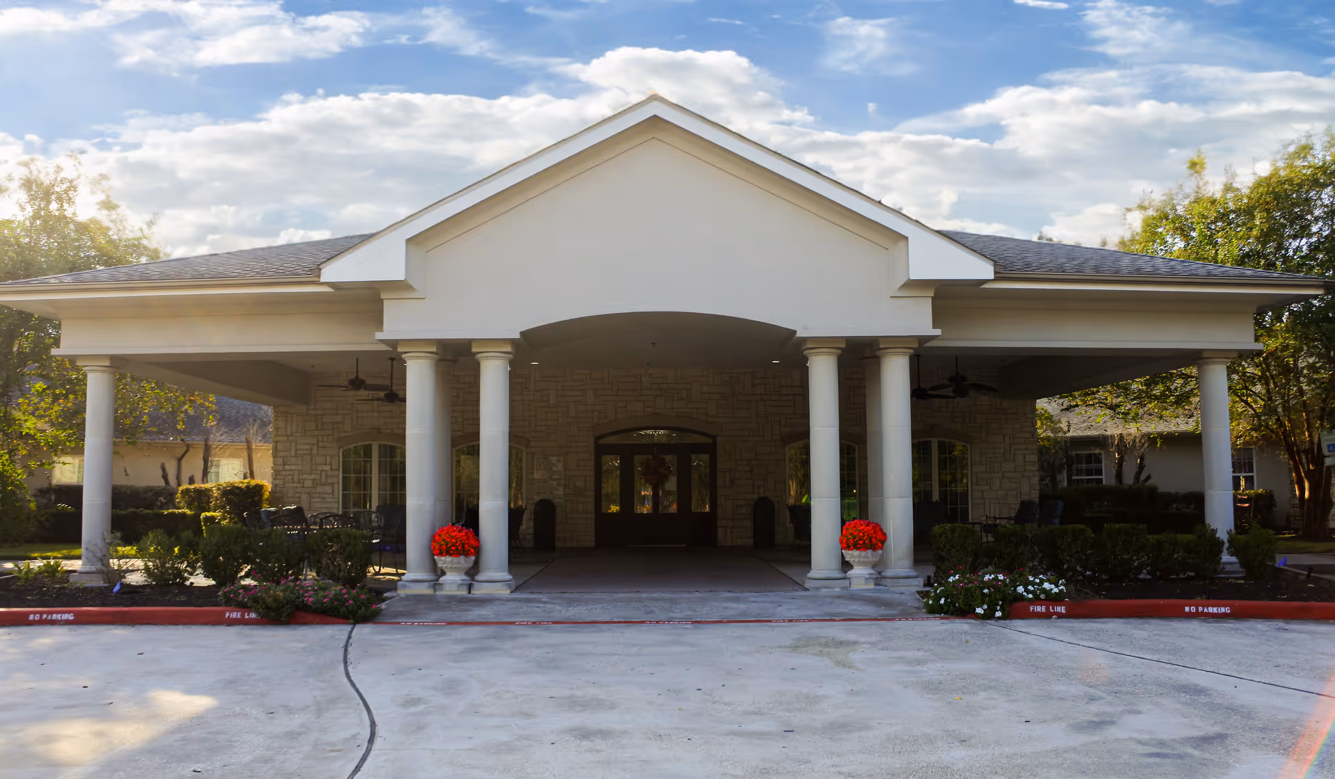 Front exterior view of Kingwood Rehabilitation & Healthcare Center featuring a covered entrance with white columns, stone walls, and a driveway with red curbs marked no parking and fire line. There are potted red flowers on either side of the entrance and some greenery around the building.