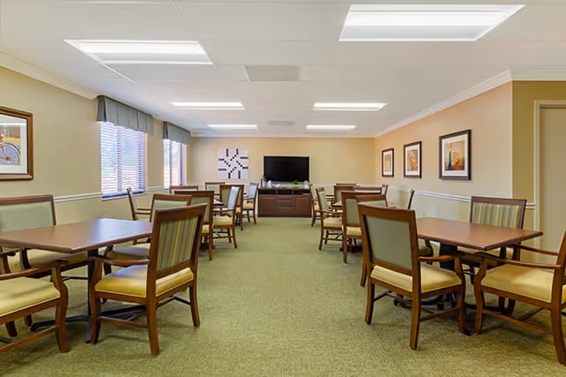 A well-lit dining room with multiple wooden tables and chairs arranged neatly. The room has large windows with blinds on the left side, framed artwork on the walls, and a television mounted on a cabinet at the far end.