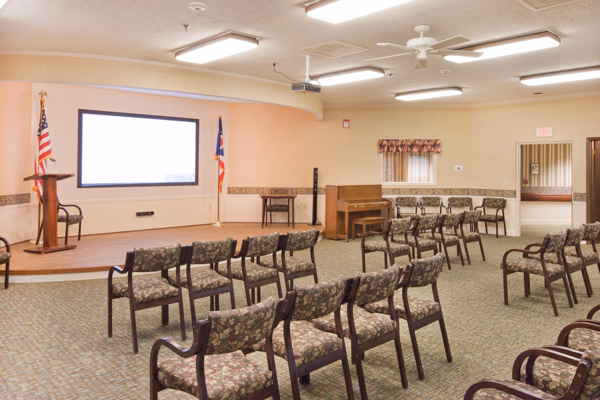 Empty activity room with rows of floral-upholstered chairs facing a small stage with a podium, projection screen, flags, and a piano.