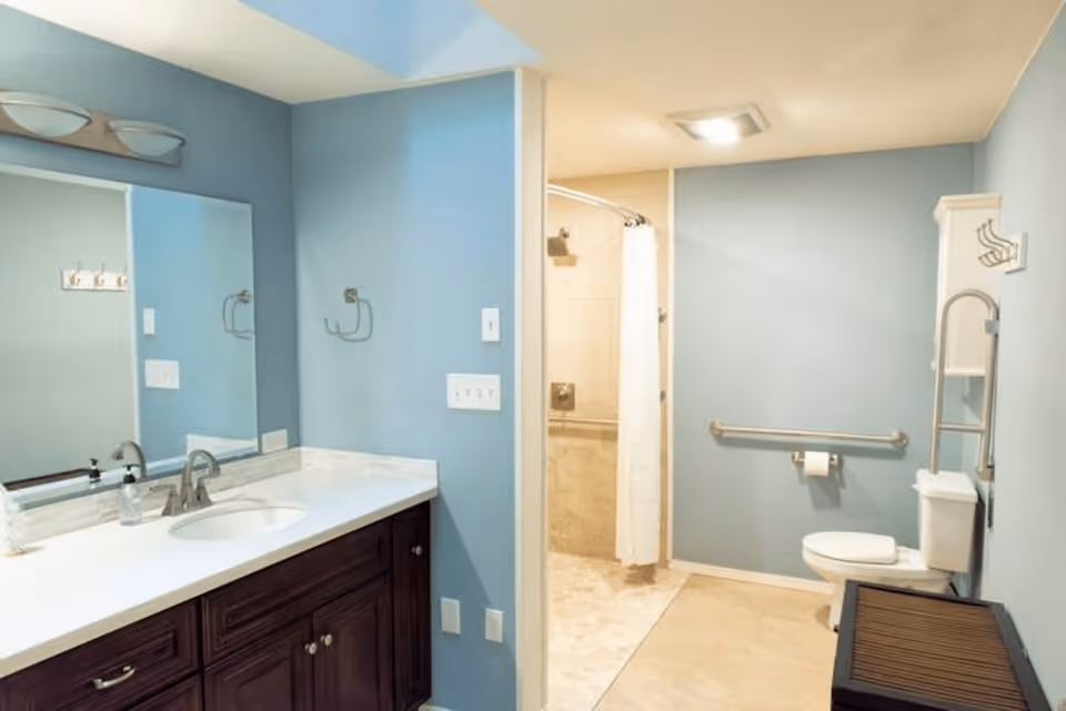 A bathroom with light blue walls featuring a white countertop with a sink and faucet on a dark wooden cabinet. There is a large mirror above the sink with two light fixtures. The bathroom includes a toilet with grab bars and a walk-in shower with a white curtain.