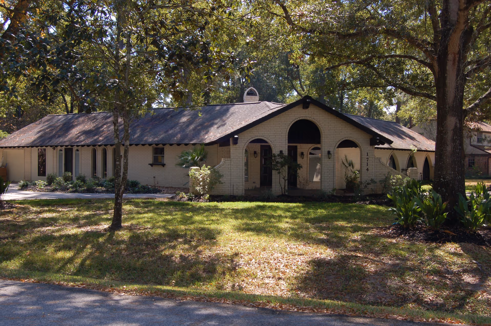Exterior view of a single-story brick building with arched windows and doorways, surrounded by trees and greenery on a sunny day.