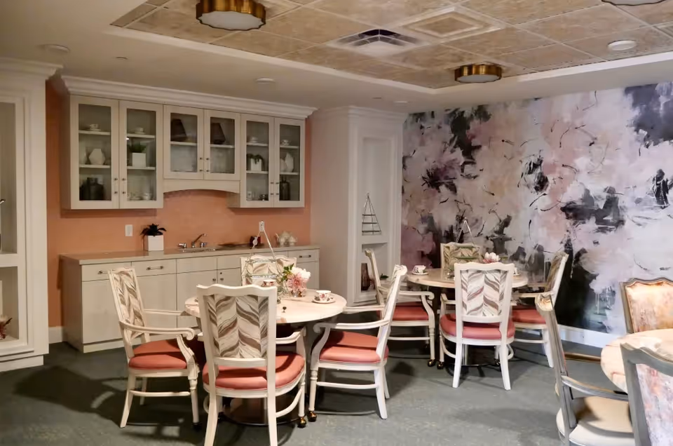 A dining room with round tables and cushioned chairs arranged around them. The walls feature a large floral mural and built-in cabinets with glass doors displaying decorative items. The room has a soft, warm lighting and a cozy atmosphere.
