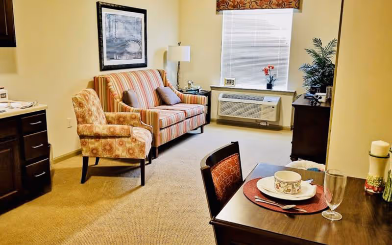 A cozy living area with a striped loveseat, a patterned armchair, a wooden dining table set with a plate, bowl, glass, and utensils, a window with blinds and a floral valance, a floor lamp, a framed picture on the wall, and a wooden cabinet with a plant on top.