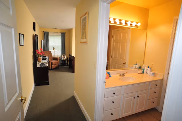 View down a hallway with beige walls and carpeted floor leading to a living area with an armchair, side table, and window with curtains. To the right, there is a bathroom vanity with a large mirror, countertop sink, and several bottles of toiletries under bright vanity lights.