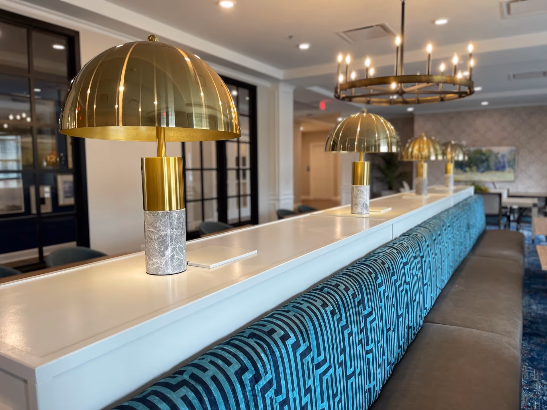 Modern lounge area with a long white counter lined with gold dome lamps and teal patterned bench seating.
