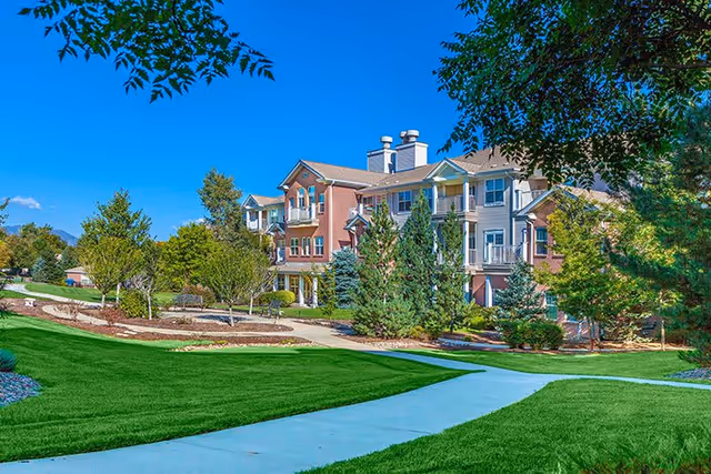 Exterior view of a multi-story senior living facility building surrounded by well-maintained green lawns, trees, and shrubs under a clear blue sky.