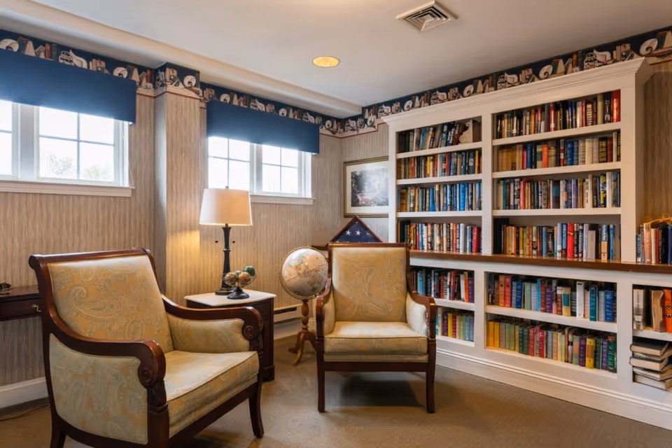 A cozy reading nook with two upholstered armchairs, a small side table with a lamp, a globe on a stand, and a built-in bookshelf filled with books. The room has two windows with blue valances and wallpaper with a decorative border near the ceiling.