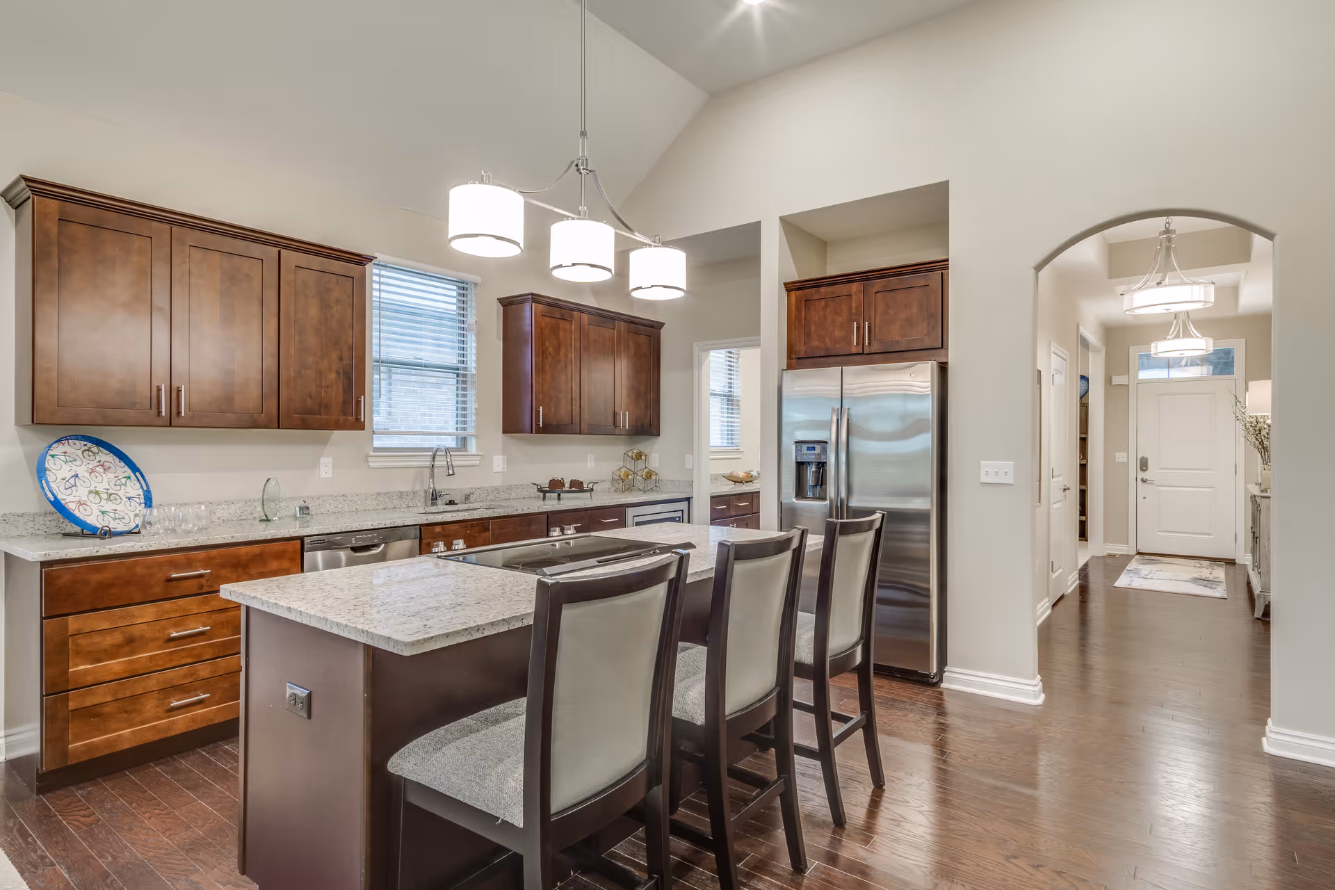 A modern kitchen with dark wood cabinets, a granite countertop island with three cushioned chairs, stainless steel refrigerator, and a window above the sink. The kitchen has hardwood floors and is well-lit with ceiling lights. An arched doorway leads to a hallway with a front door visible in the background.