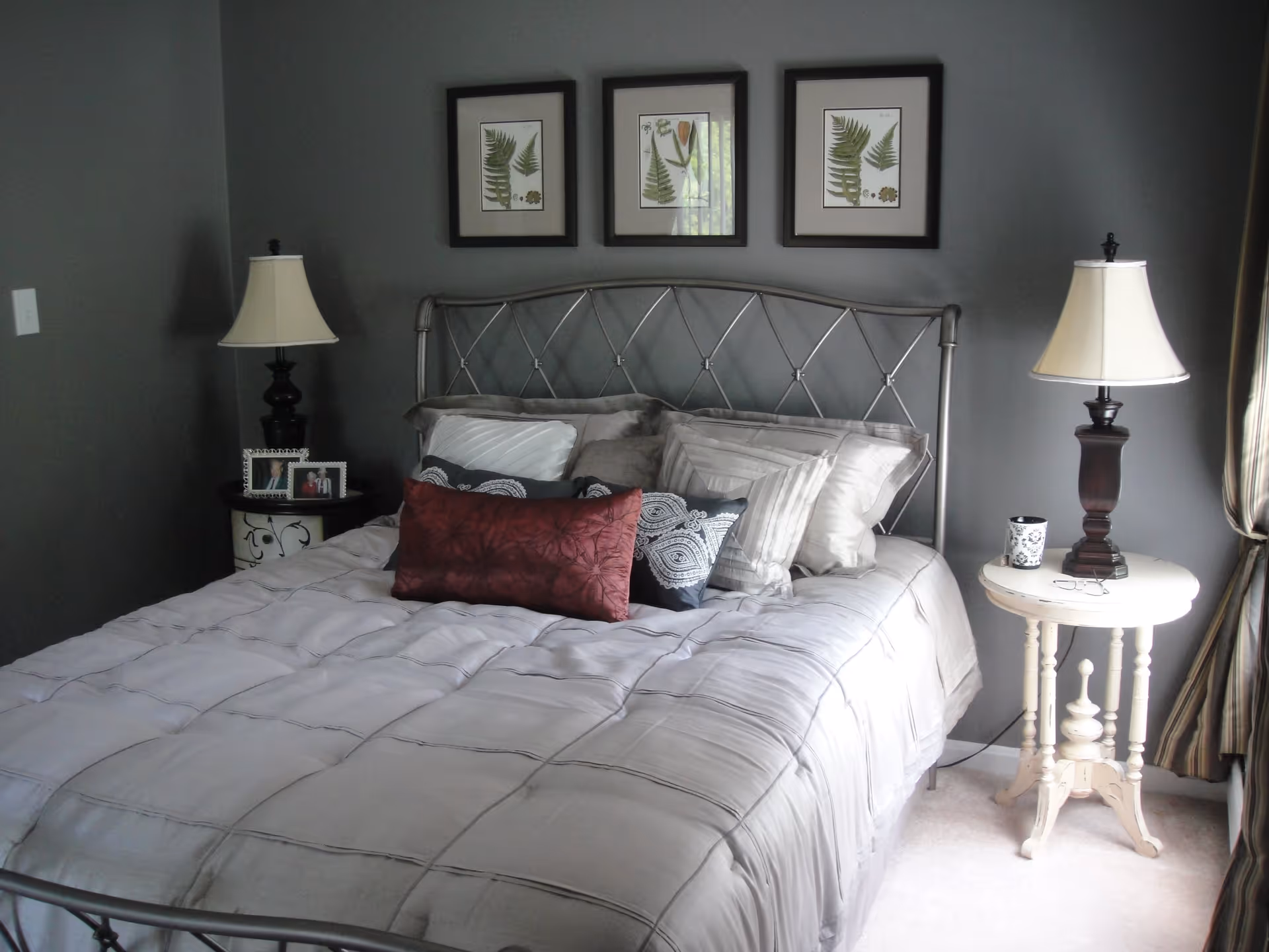 A neatly made bed with a metal headboard in a bedroom with gray walls. The bed has multiple pillows in shades of gray, black, and red. On either side of the bed are bedside tables with lamps, one black and one white. Above the bed are three framed botanical prints featuring green leaves.