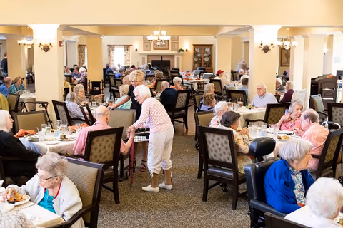 A large dining room filled with elderly residents sitting at tables eating and socializing. A woman in a striped shirt is standing and interacting with a seated man. The room has warm lighting, carpeted floors, and traditional decor including chandeliers and framed pictures on the walls.