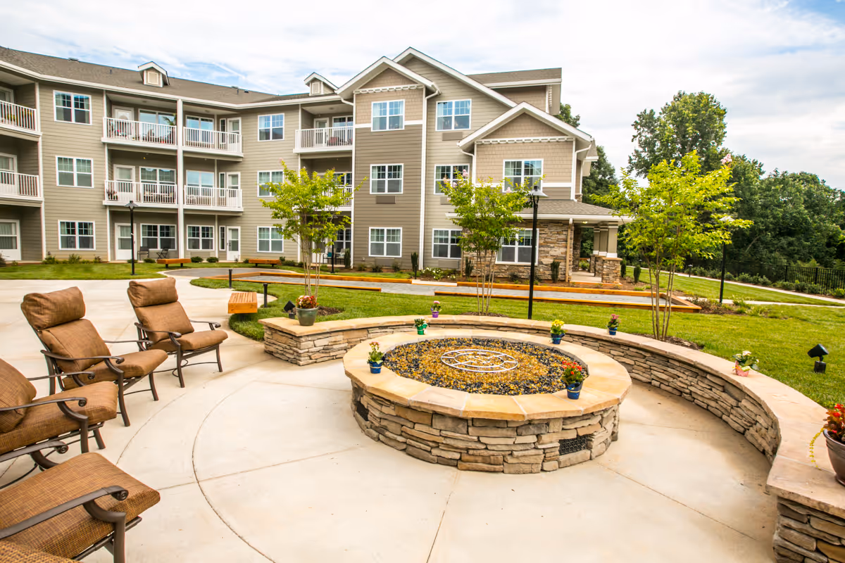 Outdoor seating area with cushioned chairs arranged around a circular stone fire pit, surrounded by a curved stone bench. In the background, there is a three-story residential building with balconies and windows, green grass, small trees, and a partly cloudy sky.