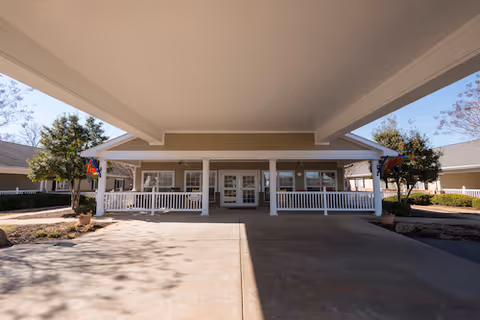 Covered drive-up entrance with white railings and double doors at the front of a single-story building flanked by potted plants and trees.