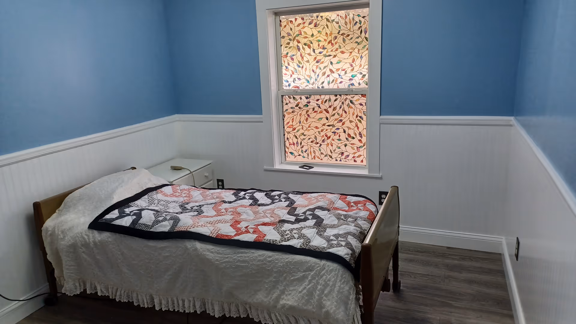 A small bedroom with light blue walls and white wainscoting. There is a single bed with a white bedspread and a colorful quilt on top. Next to the bed is a white nightstand with a lamp. A window with decorative stained glass featuring a leaf pattern is centered on the wall behind the bed.