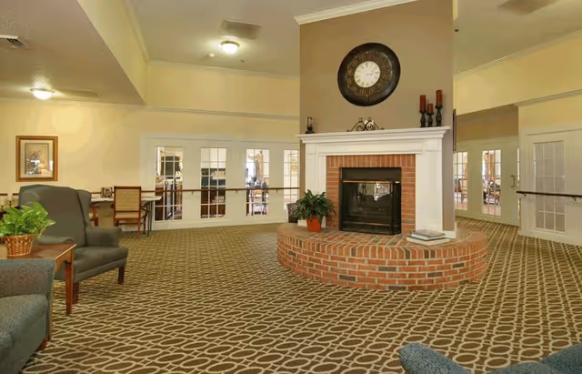 A spacious common area with patterned carpet, a central brick fireplace with a white mantle, a large round clock above the fireplace, and several chairs and tables arranged around the room. The walls are light-colored, and there are glass-paned double doors leading to another room.