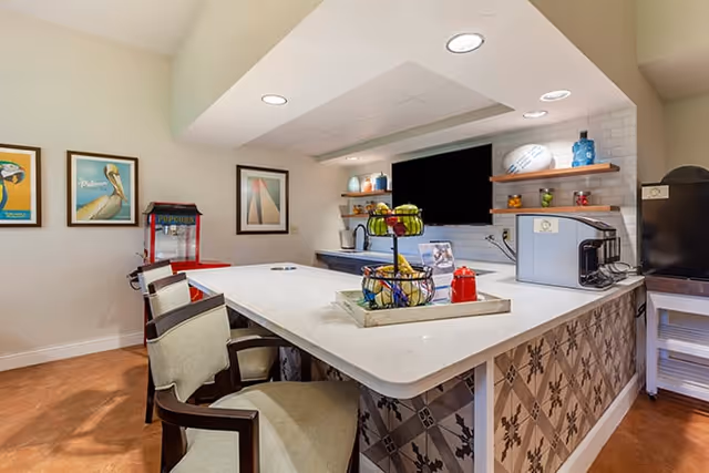 Interior view of a kitchen area in a senior living facility featuring a large white countertop island with patterned tile on the side, two chairs, a popcorn machine, framed artwork on the wall, shelves with decorative items, a large flat-screen TV, and kitchen appliances including a coffee maker.