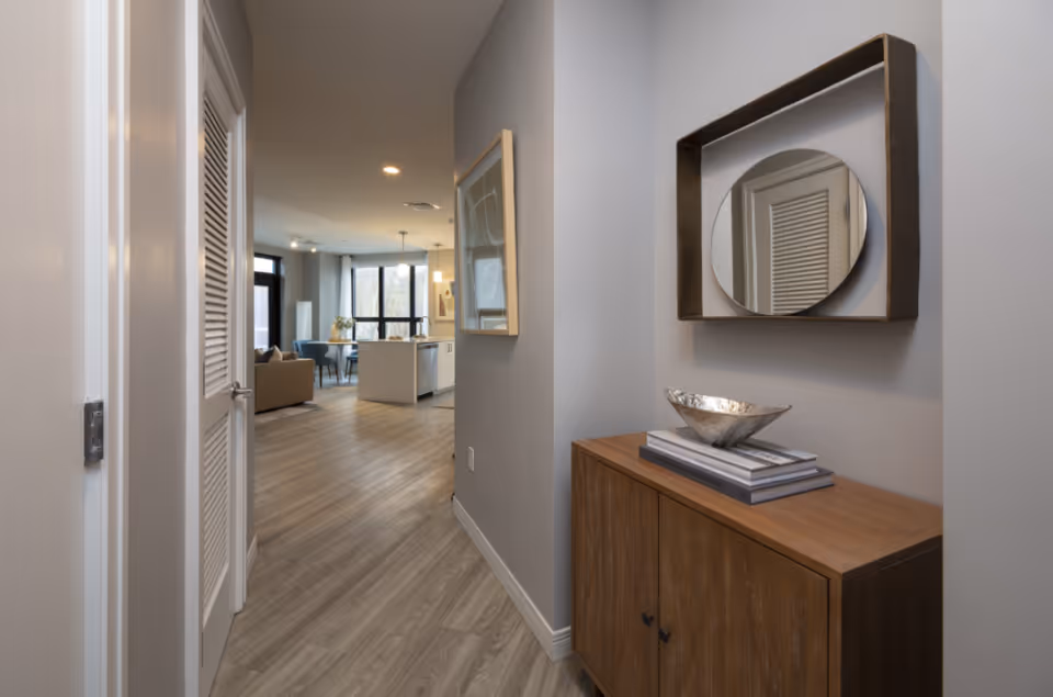 Hallway with a wooden console and round mirror leading into an open-plan living room and kitchen with wood flooring.