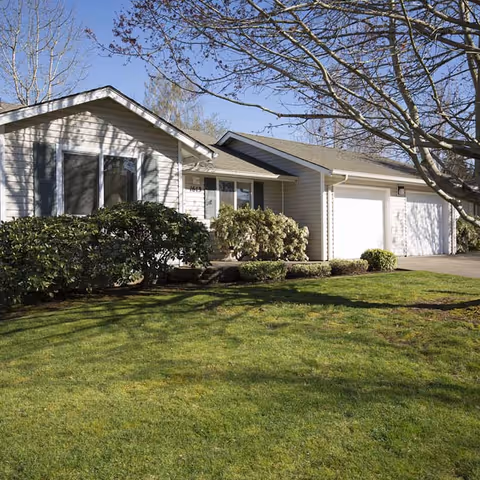 Front exterior of a single-story house with an attached garage, manicured lawn, and shrubs under a clear blue sky.