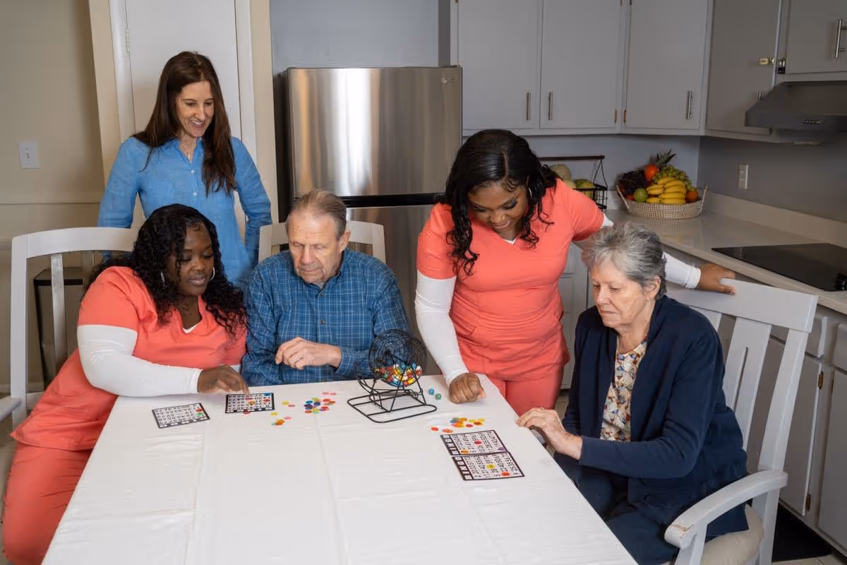 Two elderly individuals and two caregivers playing bingo at a table in a kitchen area, with a woman standing and watching them. The kitchen has white cabinets, a stainless steel refrigerator, and a basket of fruit on the counter.