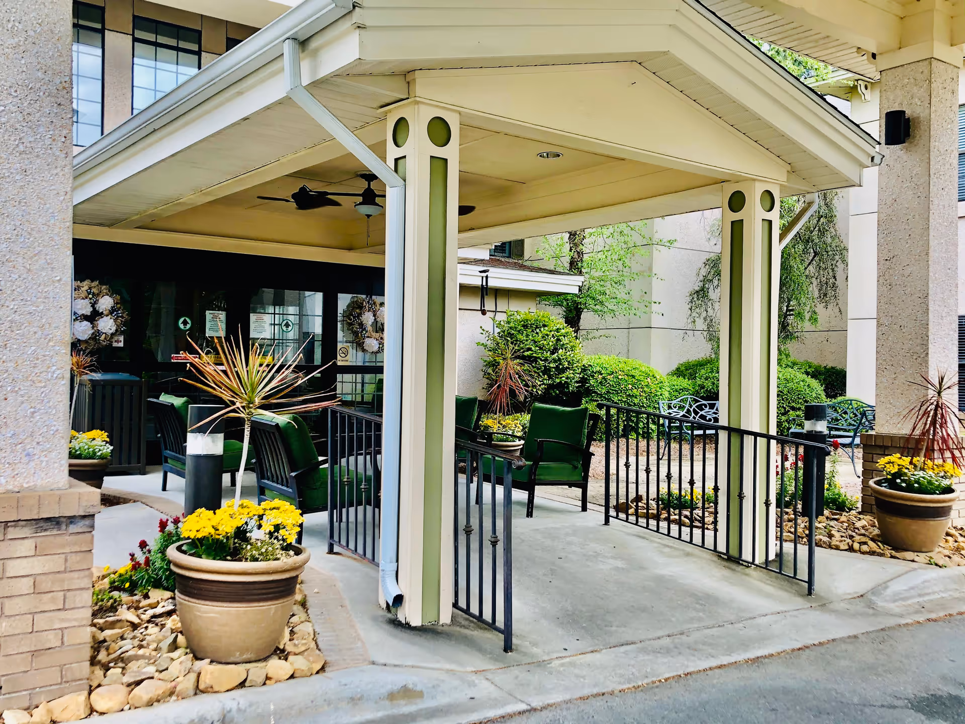 Covered outdoor seating area at the entrance of a building with green cushioned chairs, potted plants with yellow flowers, and decorative bushes in the background.