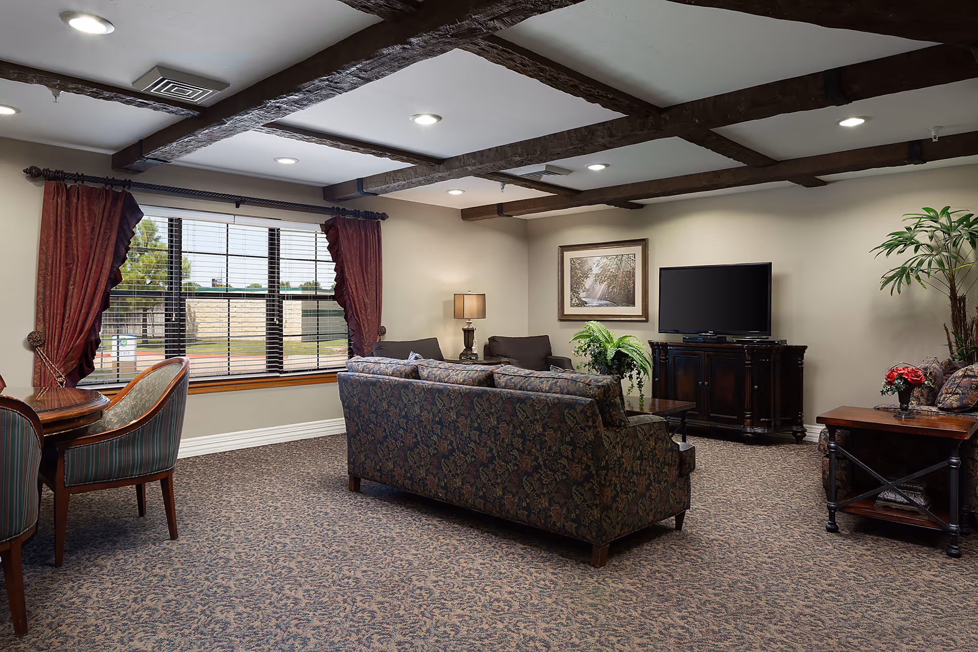 A cozy communal living room with patterned sofas, a TV on a wooden cabinet, a window with red curtains, and exposed ceiling beams.