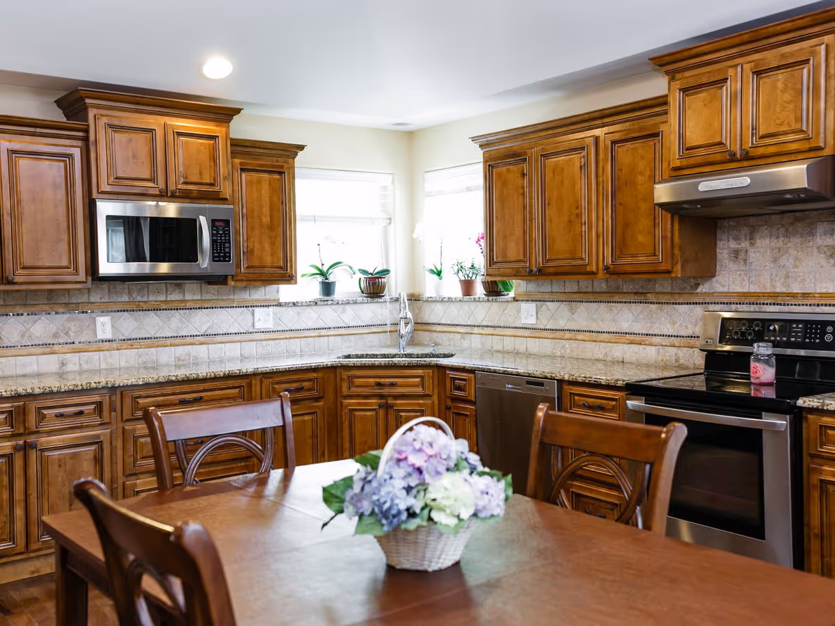 A kitchen with wooden cabinets, granite countertops, a stainless steel microwave, stove, and dishwasher. There are two windows with potted plants on the windowsill. In the foreground, there is a wooden dining table with chairs and a basket of flowers on top.