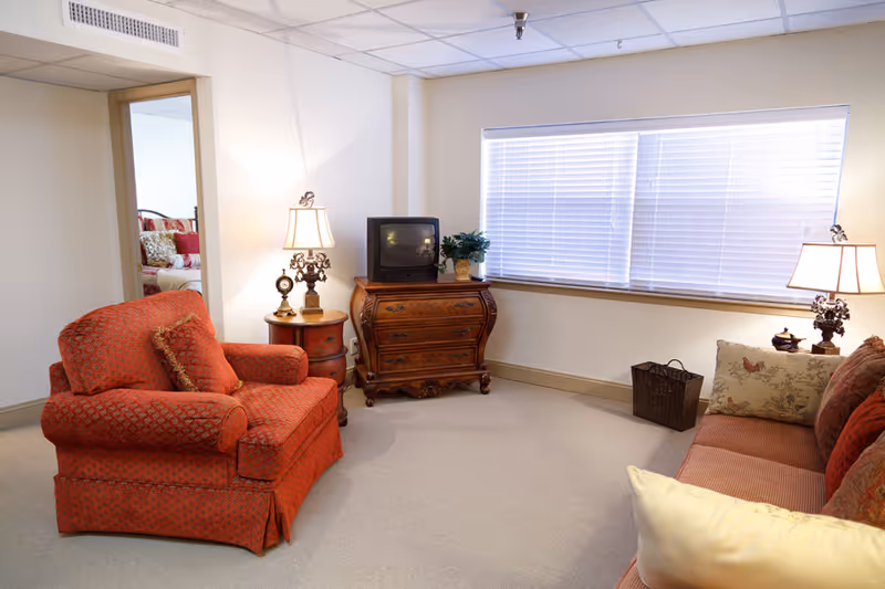 Cozy living room featuring a red upholstered armchair and matching sofa, ornate wooden TV dresser with a small television, table lamps, and a large window with blinds.