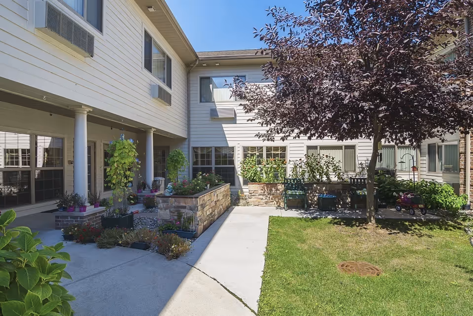 Outdoor courtyard area of a senior living facility with a concrete walkway, green grass, a tree, raised garden beds with plants and flowers, and several chairs arranged near the garden beds. The building surrounding the courtyard has white siding and multiple windows.