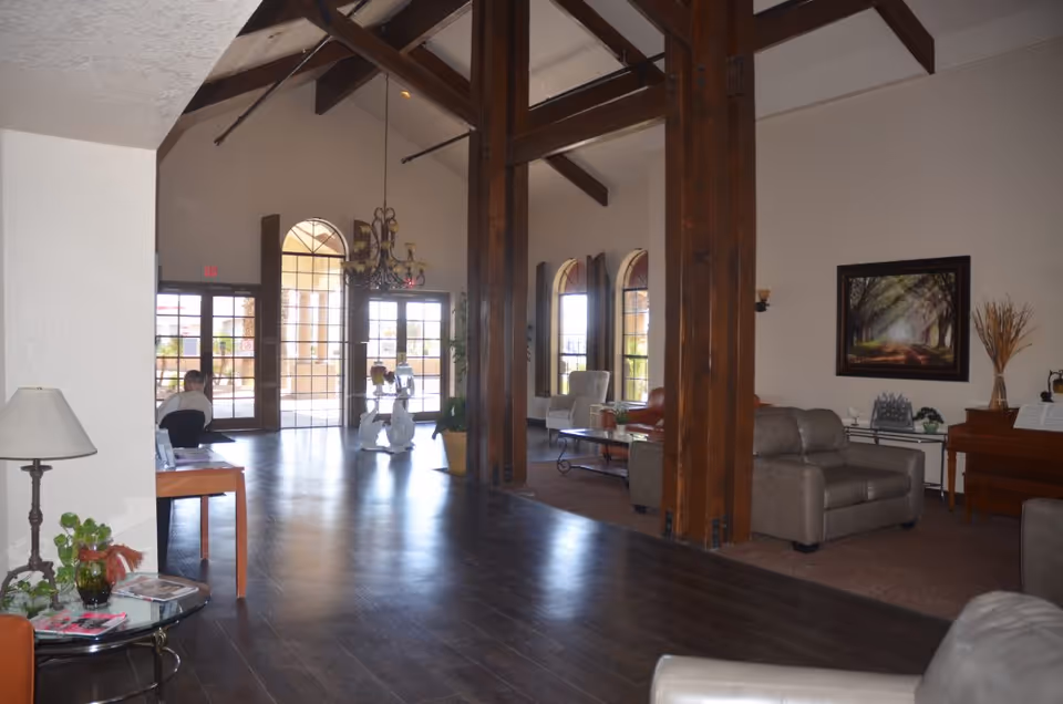 Interior view of a senior living facility common area with wooden beams, large windows, and glass doors letting in natural light. The room features comfortable seating including armchairs and sofas, a coffee table, a piano, a chandelier, and decorative plants. A person is seated at a desk near the entrance.
